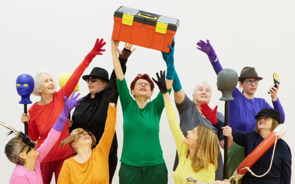 Ensemble von „Die Damen Bricolage“ in bunten Farben streckt die Arme nach einem orangefarbenen Werkzeugkasten, ringsum Requisiten.